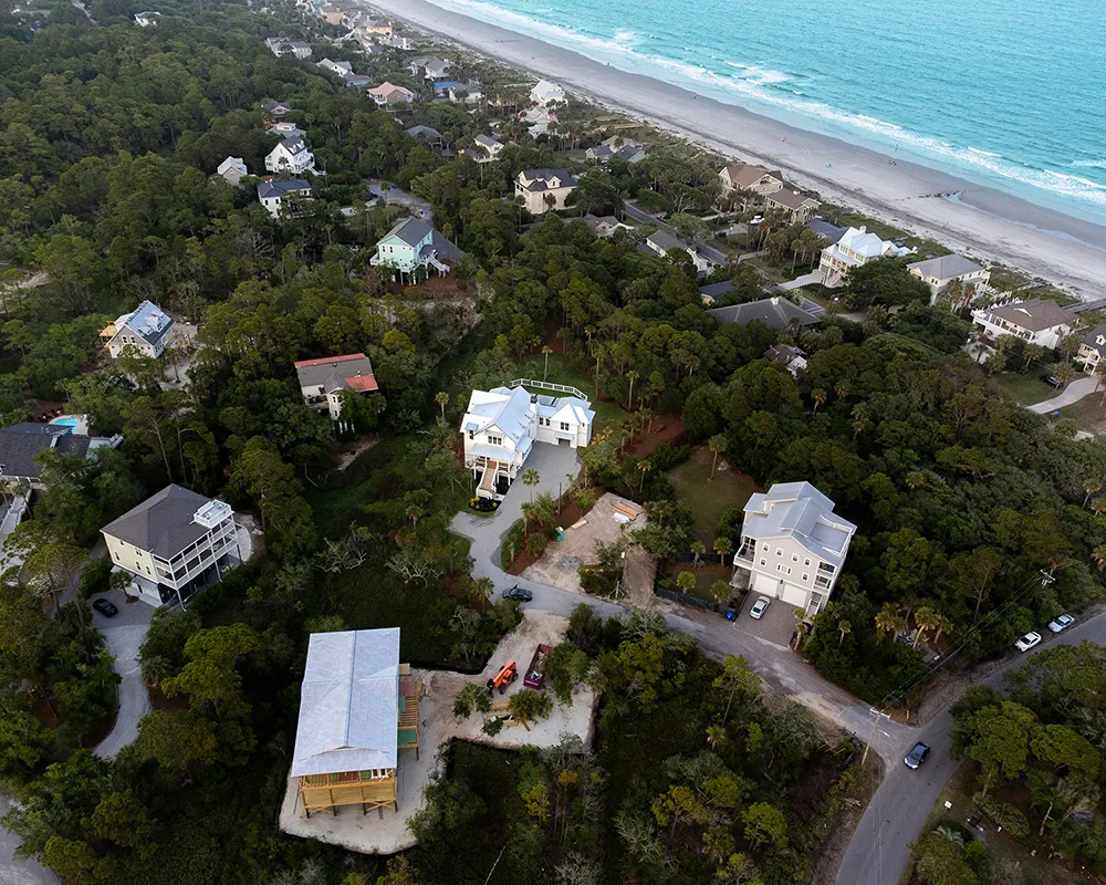 Aerial view of coastal homes surrounded by trees near a sandy beach and ocean shoreline.